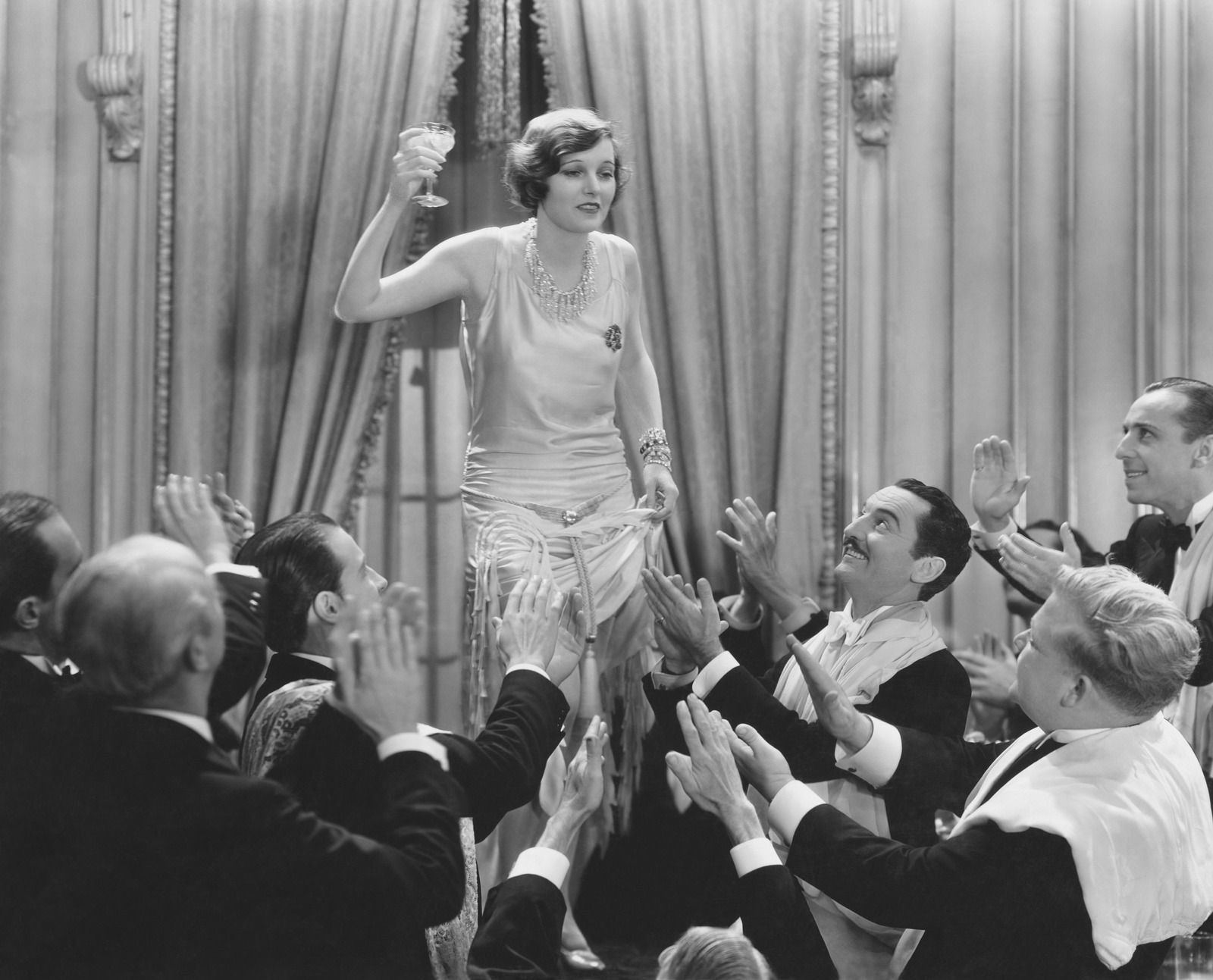 Woman in flapper dress with a champagne glass, standing atop a group of men applauding in a formal setting.