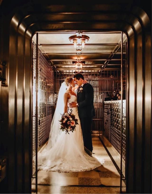 Bride and groom embrace in a dimly lit corridor with wine racks. Bride in white dress, groom in suit.