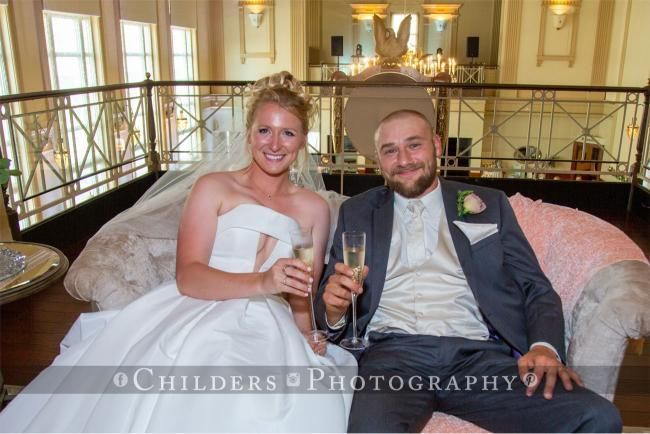 Bride and groom toast with champagne on a plush couch at a wedding.
