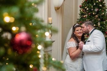 Bride and groom share a tender moment near a Christmas tree. The groom kisses the bride's cheek.