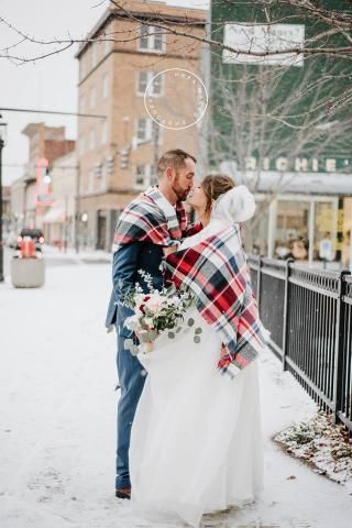 Newlyweds kissing in the snow, wrapped in a red and black plaid blanket; buildings in the background.