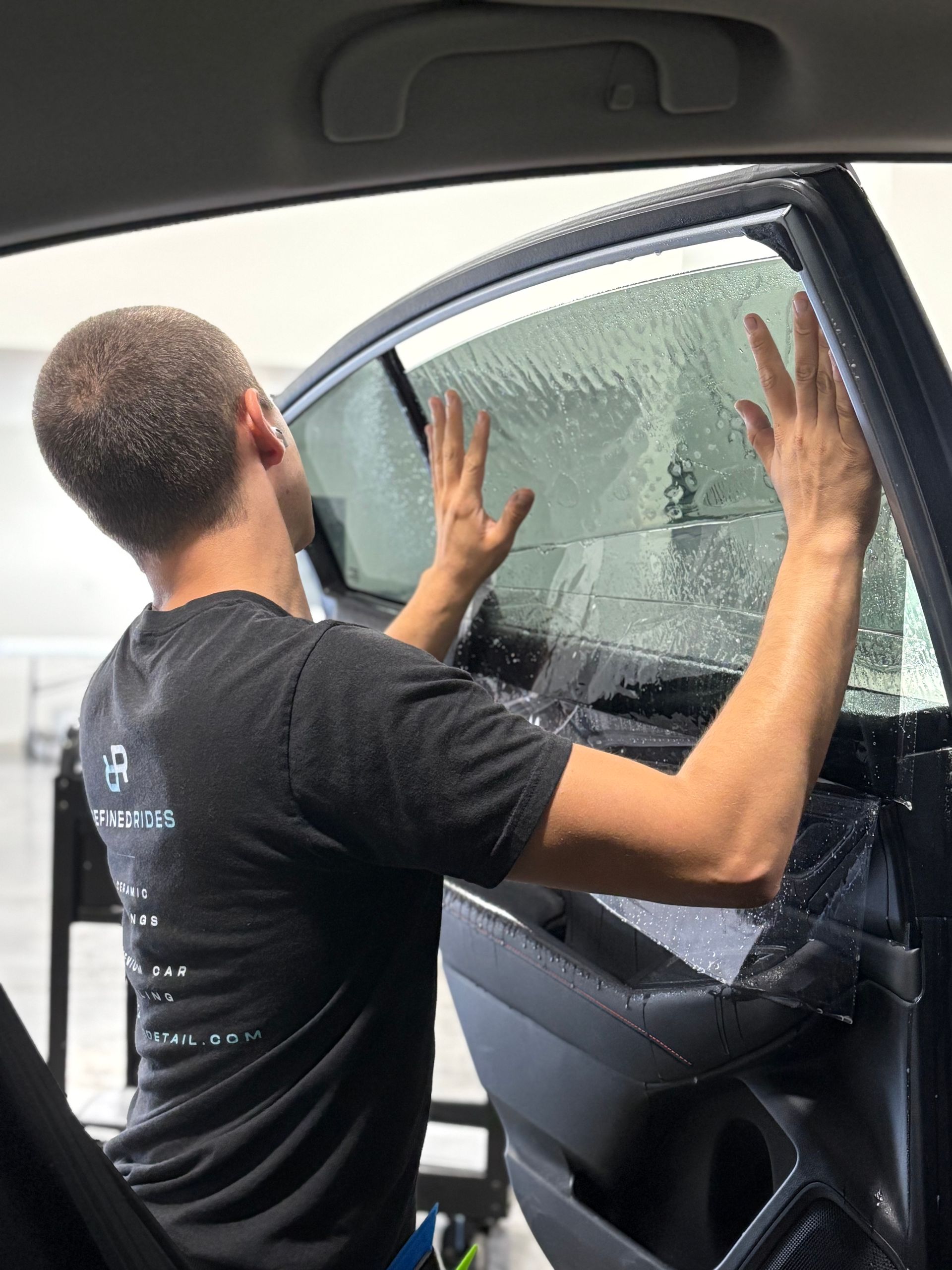 Man applying tint to car window in a garage, wearing a beanie and hoodie.