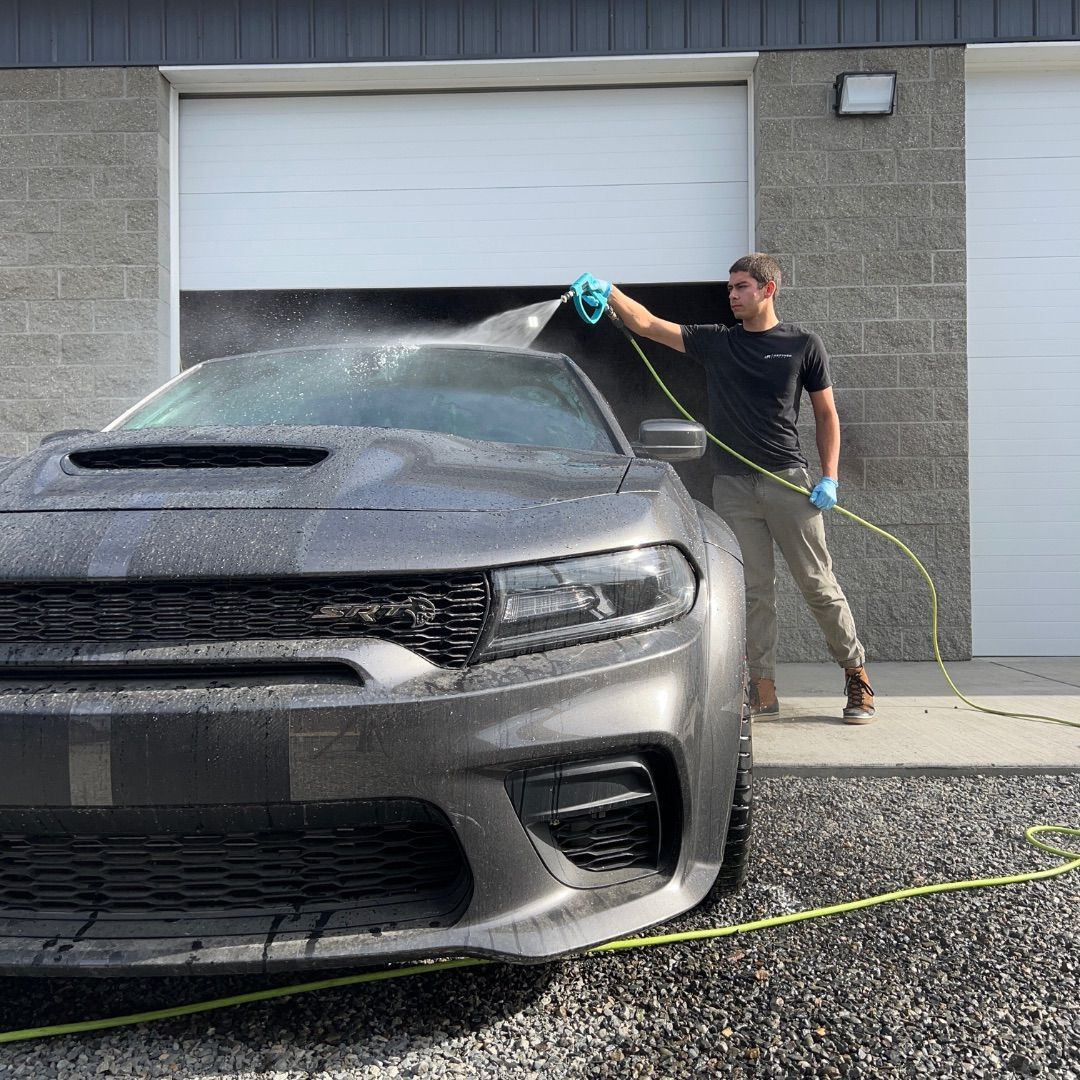 A man is washing a car with a hose in front of a garage door.