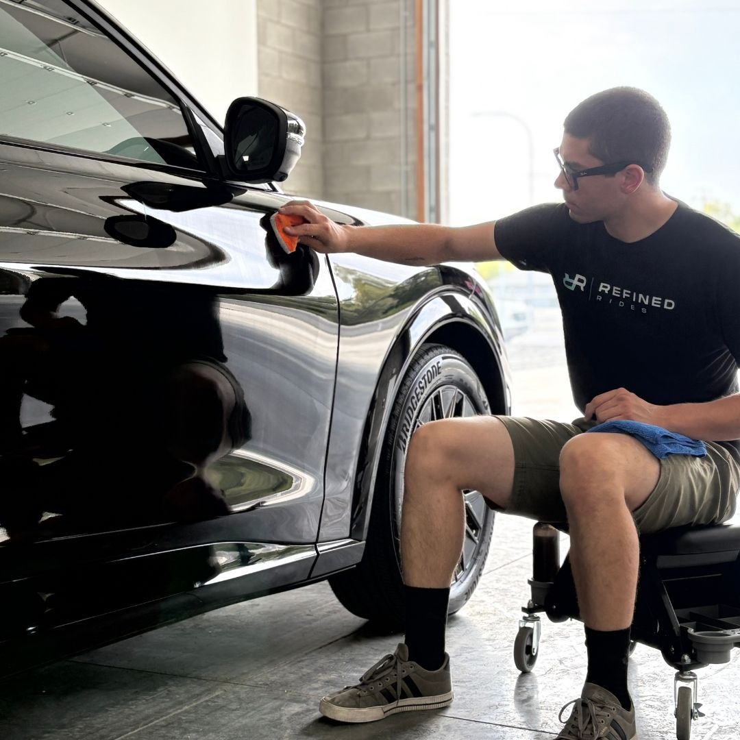 Man polishing a shiny black car, wearing a black t-shirt, shorts, and glasses.