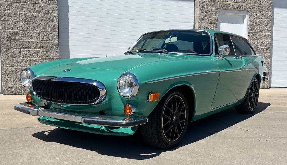 Green Volvo P1800 coupe parked in front of a building with white garage doors.