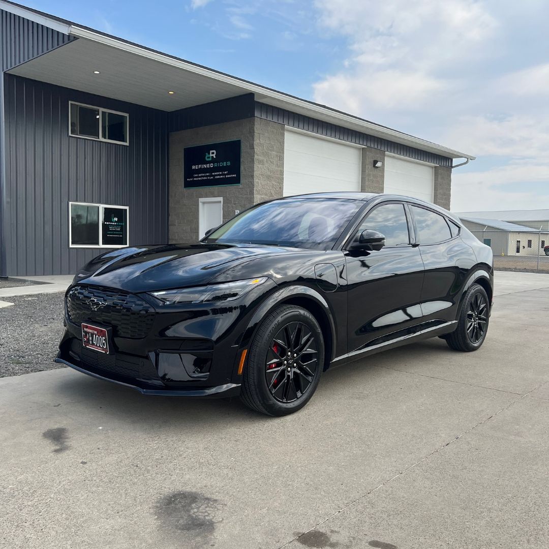 Black Ford Mustang Mach-E parked in front of a gray building with a garage door, blue sky background.