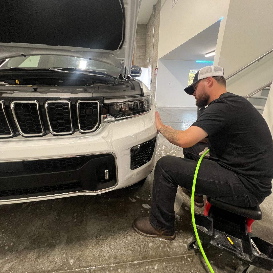 Man in black shirt and hat working on the front of a white car in a garage.