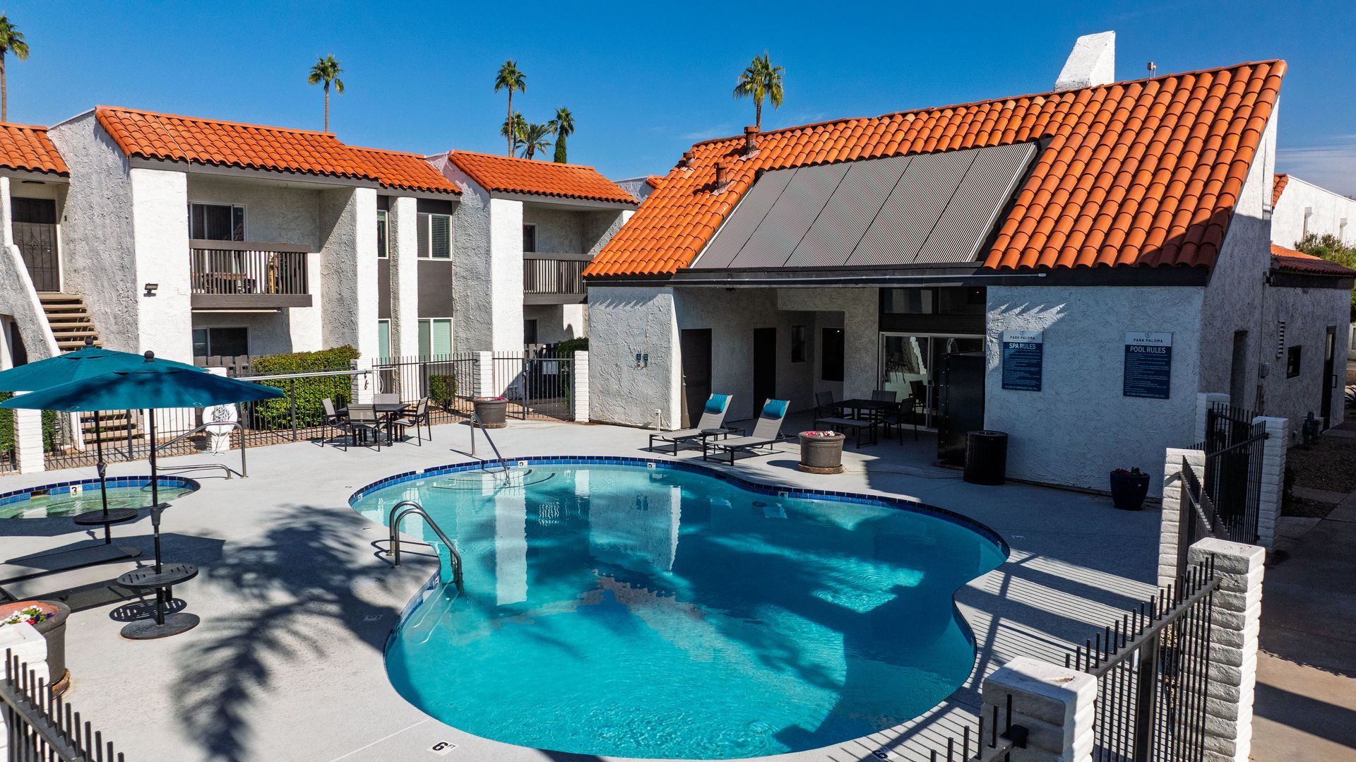 Swimming pool with lounge chairs, umbrella, and buildings with red tile roofs on a sunny day.