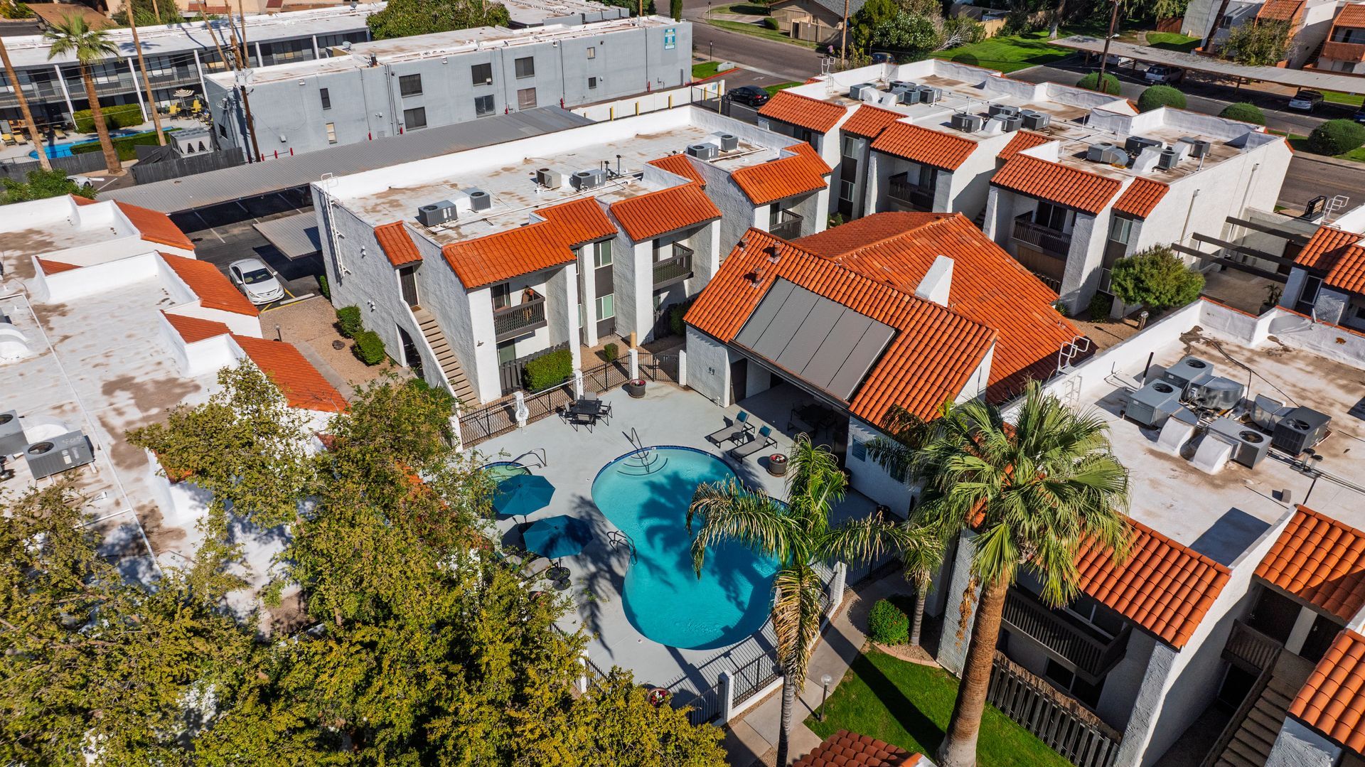 Aerial view of apartment complex with a pool, red-tiled roofs, and palm trees.