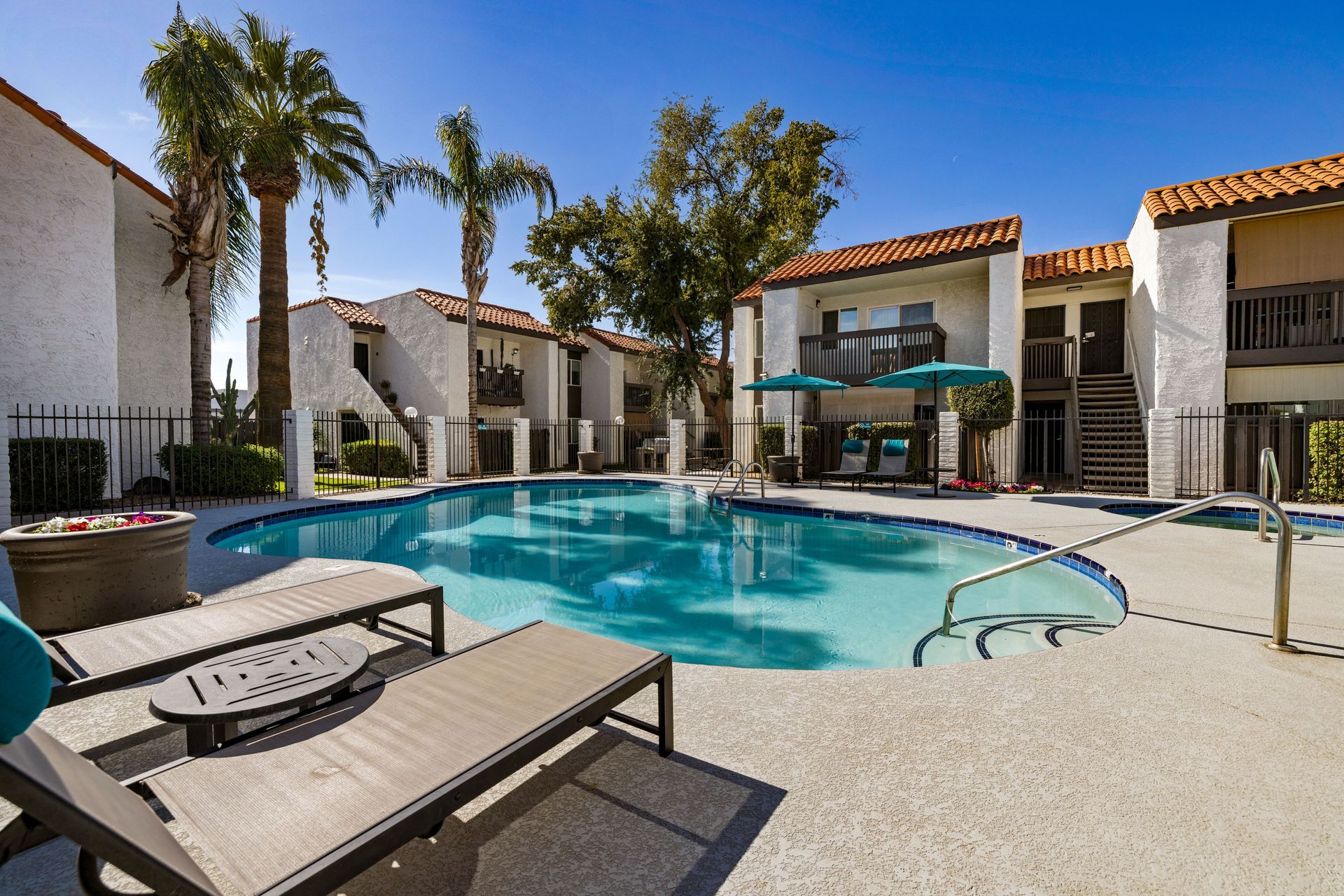 Apartment complex with a swimming pool, palm trees, and lounge chairs on a sunny day.