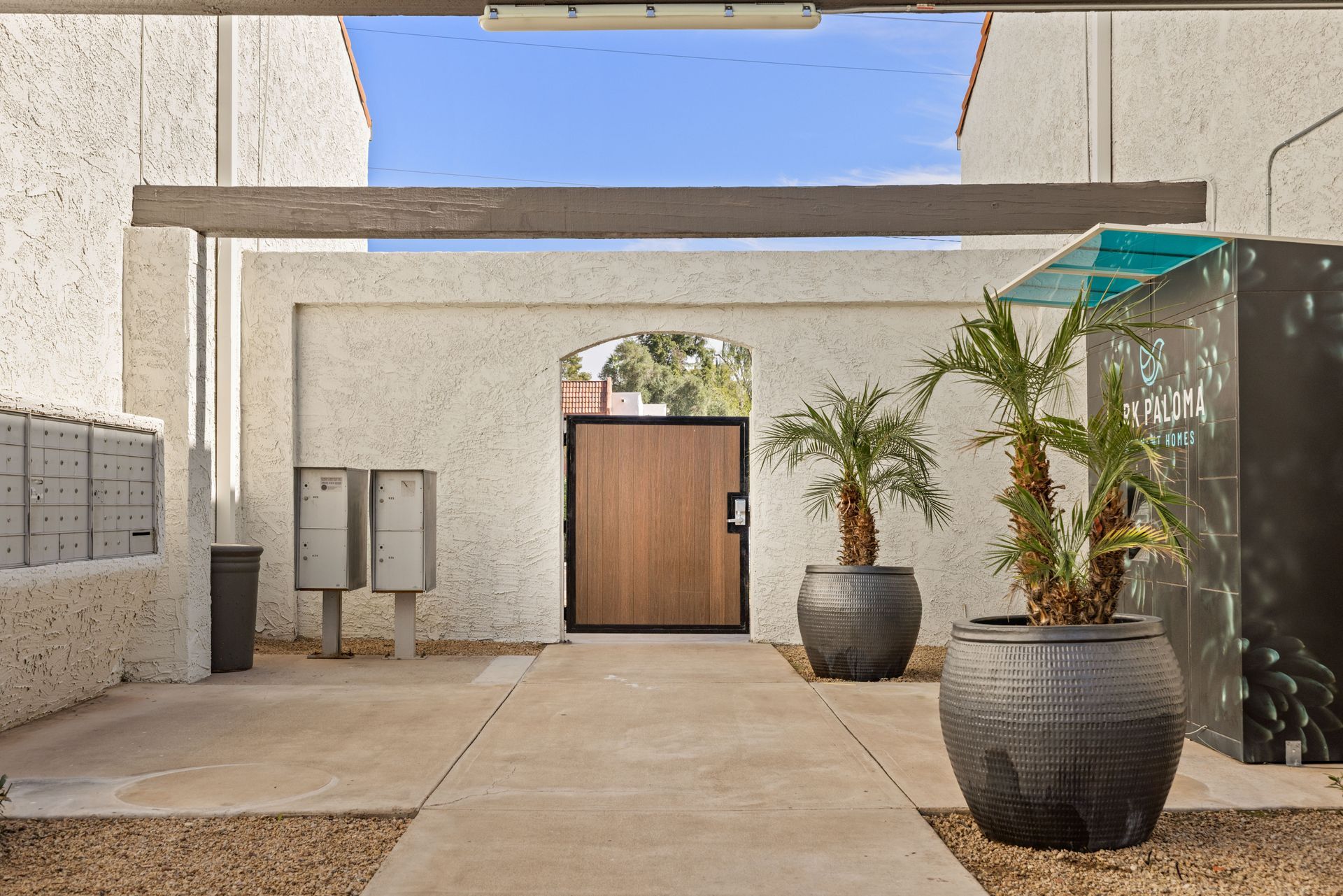 Courtyard entrance with mailboxes, wooden door, potted plants, and concrete walkway under a beam.