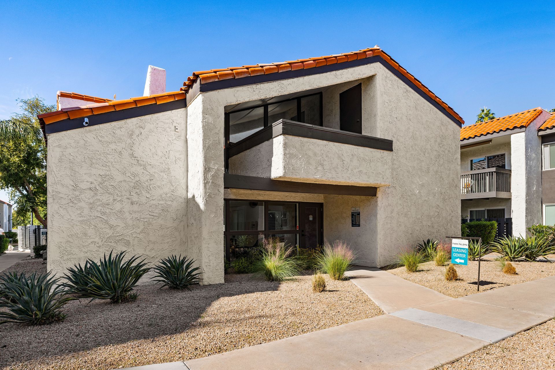 Two-story stucco apartment building with a brown roof and pathway. Blue sky and desert landscaping in front.