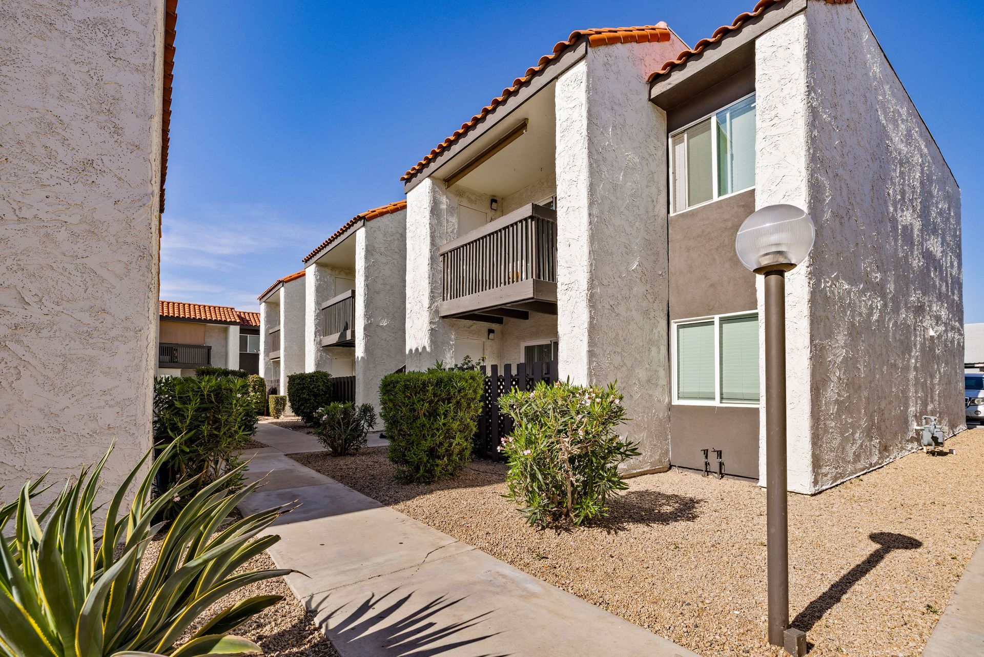 Apartment complex with stucco walls, red tile roofs, and small balconies, pathway with landscaping, bright sky.