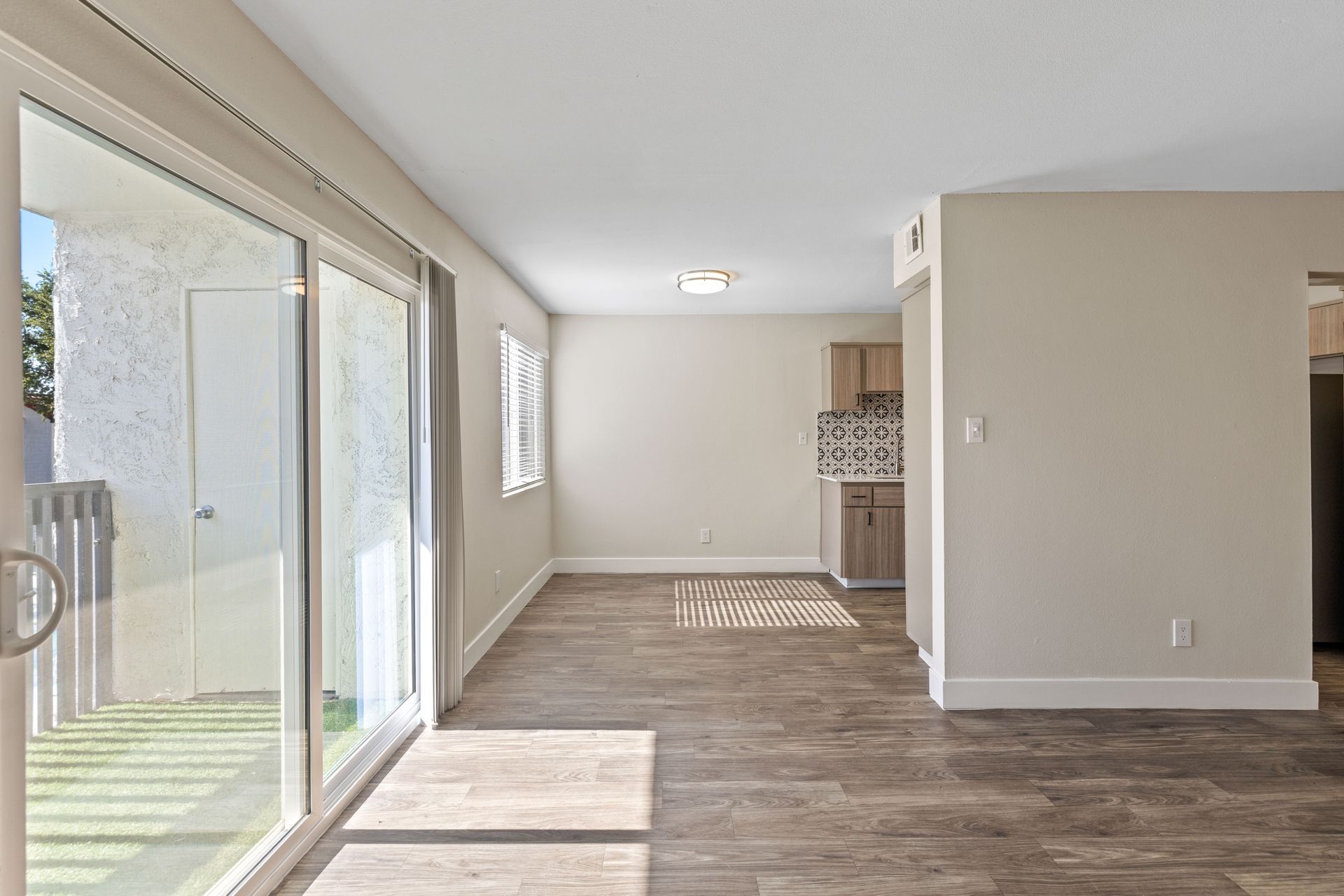 Empty living room with wood-look flooring, large sliding glass door, and a glimpse into a kitchen.