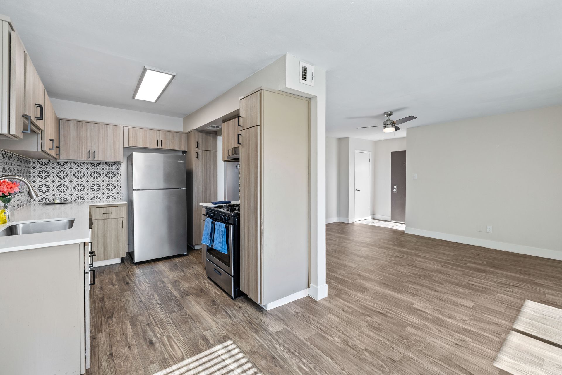 Kitchen with wood-look flooring, stainless steel appliances, light wood cabinets, and gray walls opening to a living area.