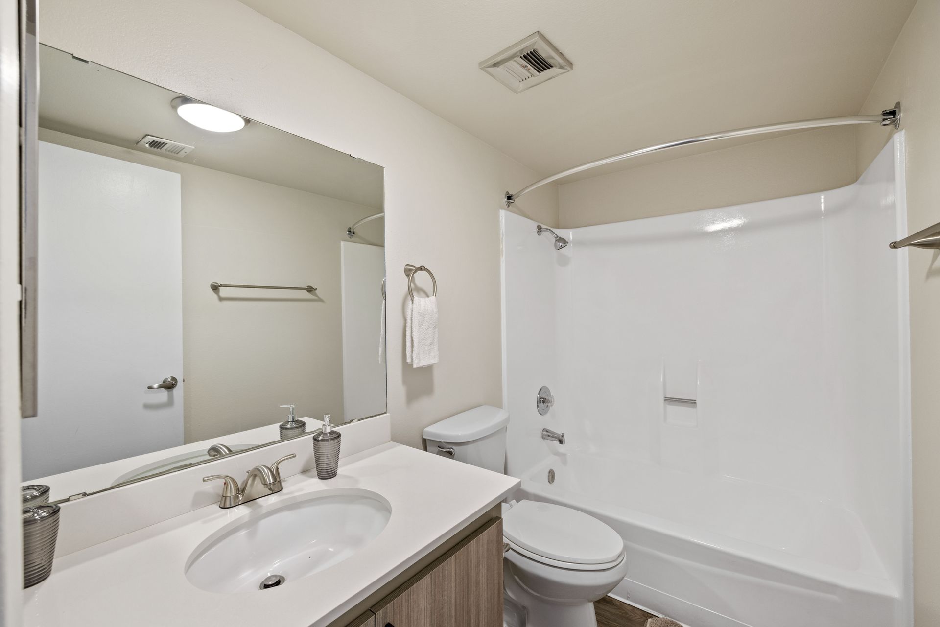 Bathroom with white tub/shower, toilet, sink, and large mirror. Neutral tones, well-lit.