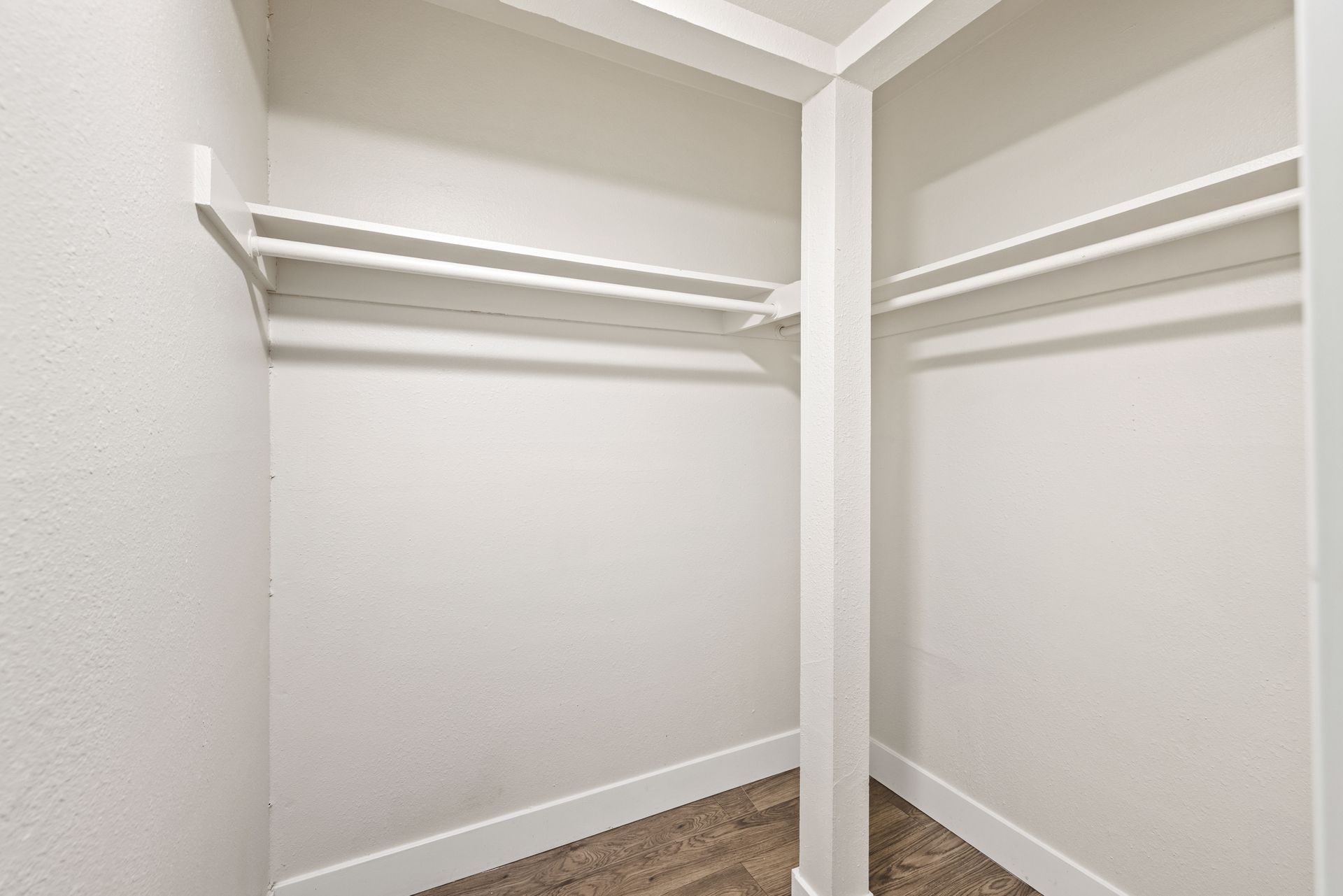 Empty white closet with hanging rods and a dividing pillar, wooden floor.