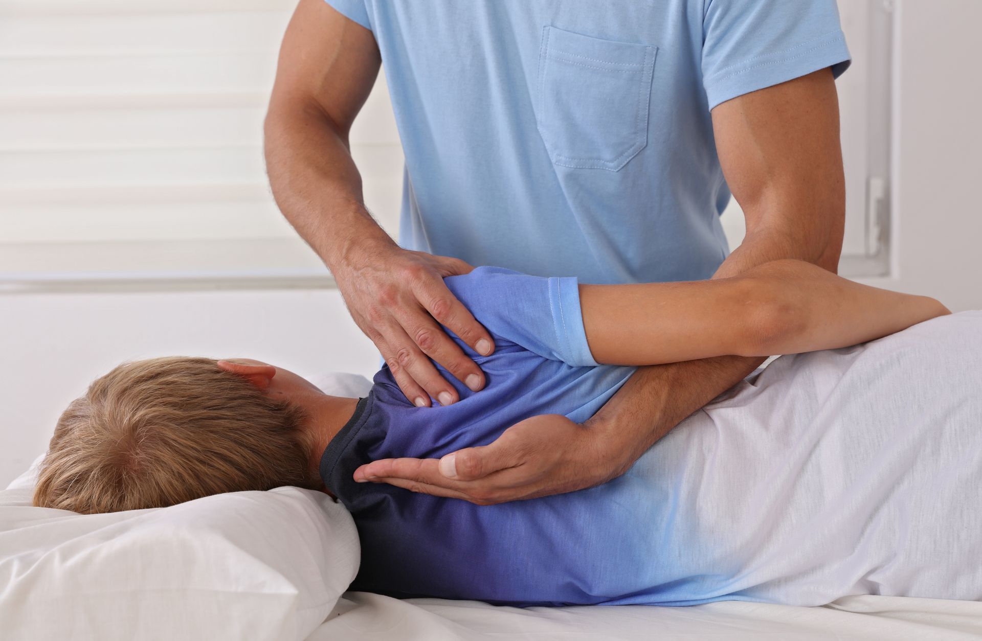 Man giving a patient a shoulder adjustment. The patient lies on a bed; the man's hands are on the patient's back and arm.