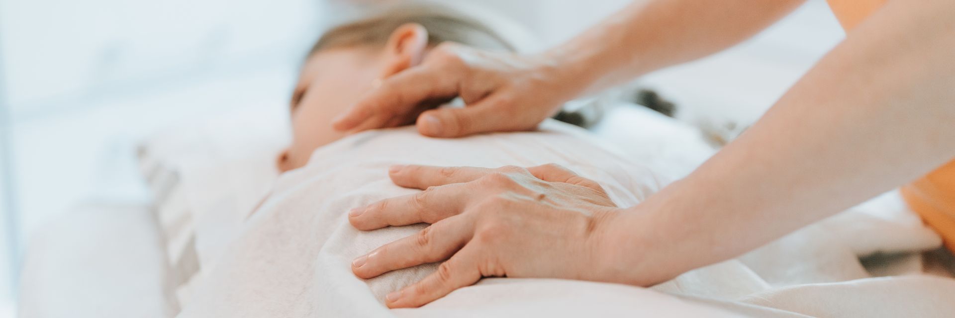 Hands of an adult giving a massage to a baby, who is lying on a white surface.