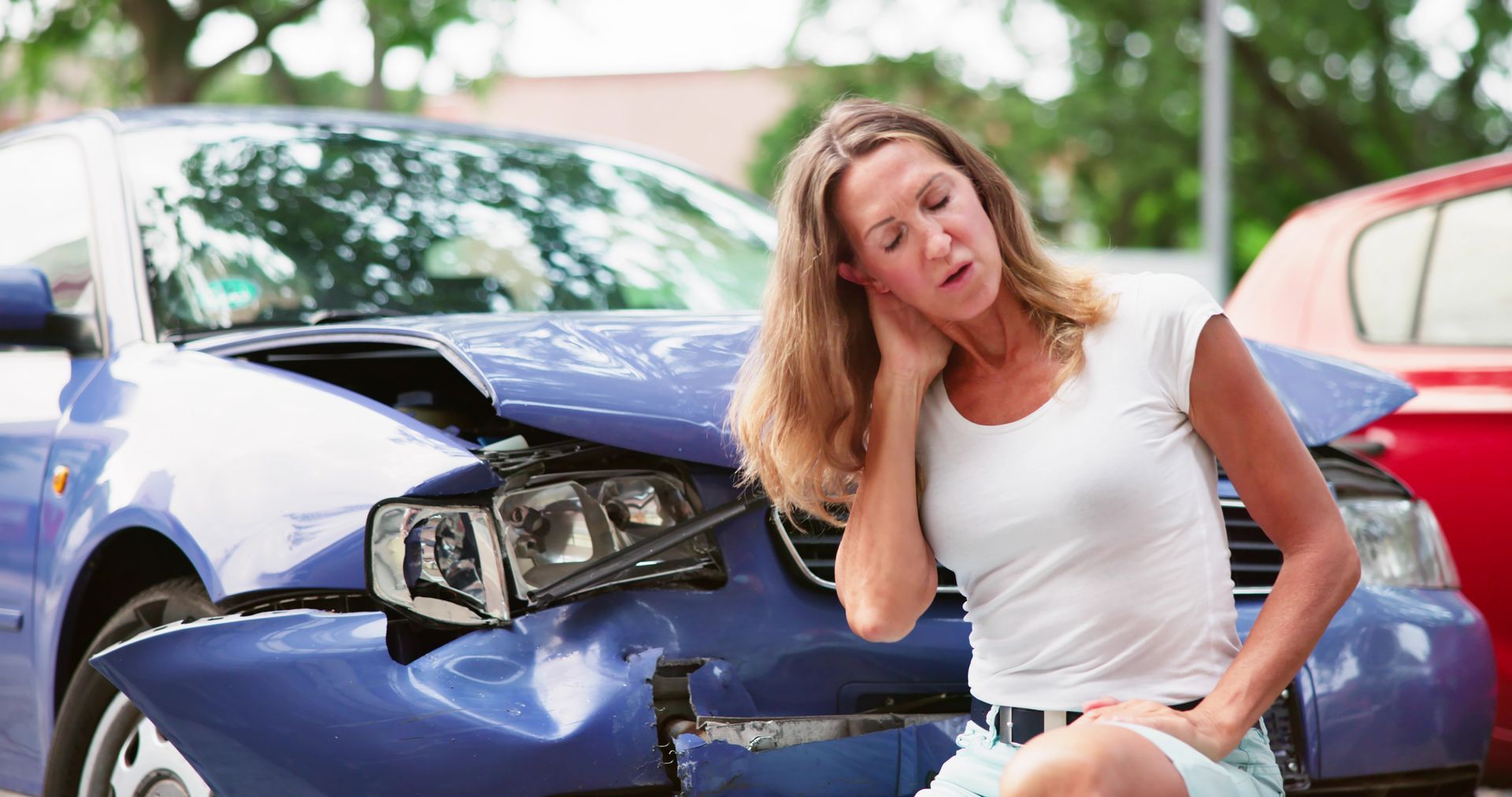 Woman holding her neck, sitting near a damaged blue car after a car accident. She appears to be in pain.