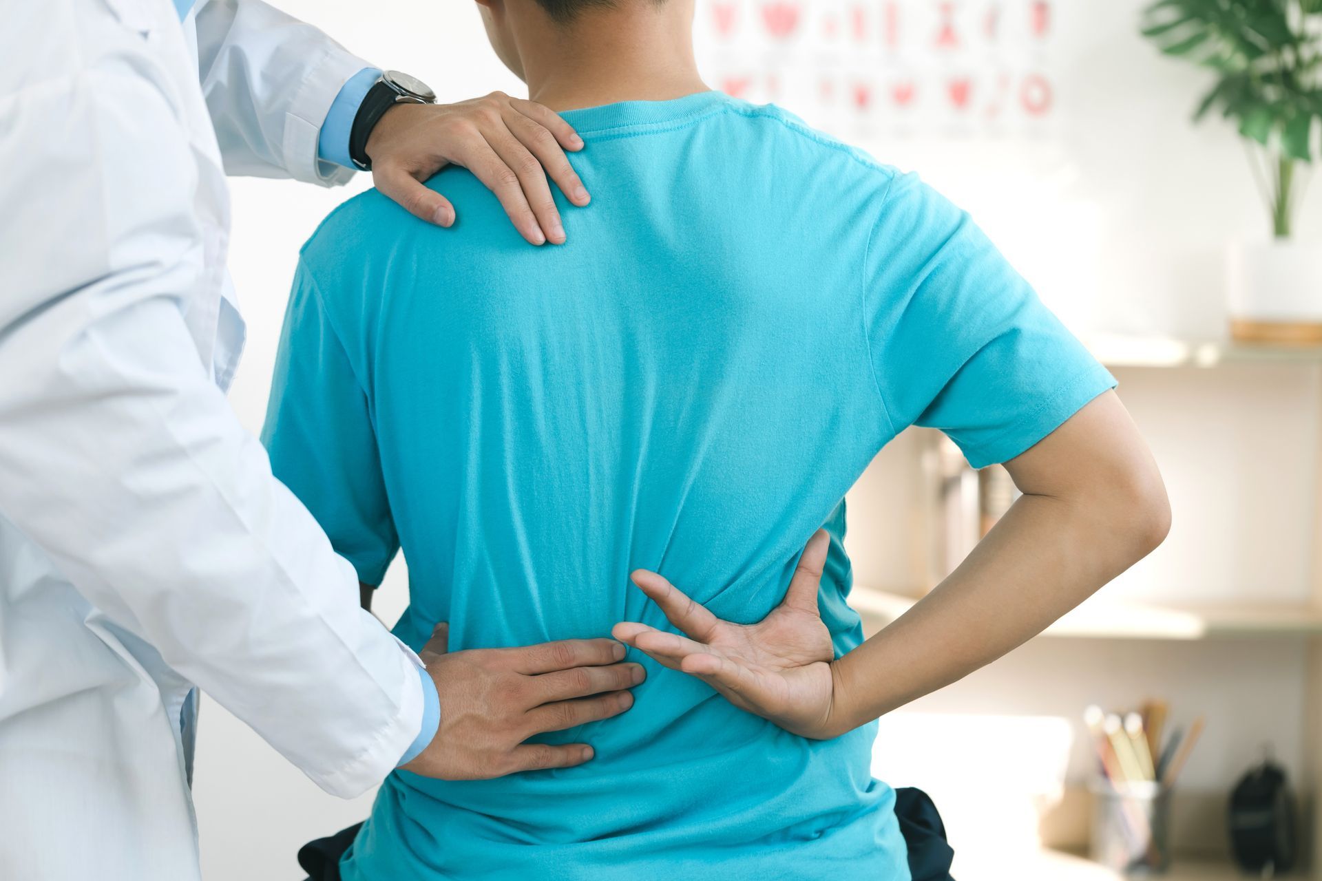 Doctor examining a patient's lower back in an office setting. The patient wears a blue shirt and is bent over, while the doctor in a white coat touches the back.