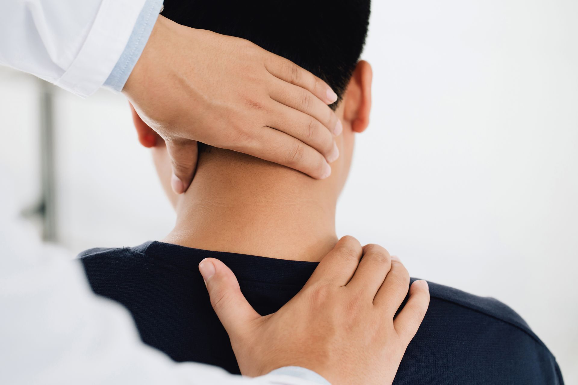 A healthcare professional examining a patient's neck. The doctor's hands are on the patient's neck and shoulder.