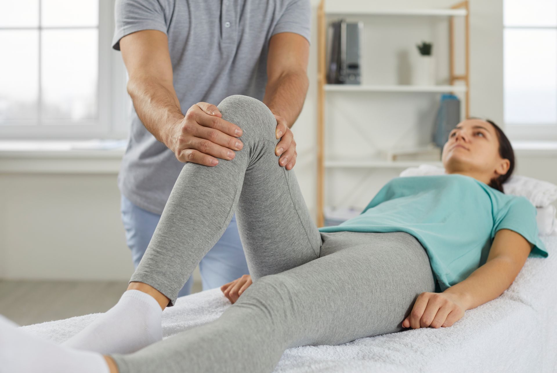 A person lies on a massage table while a therapist examines their knee in a light-filled room.