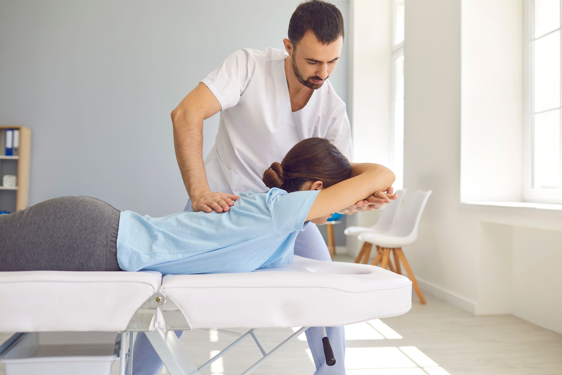 A physical therapist assisting a woman lying on a massage table, working on her shoulder and back in a light-filled room.