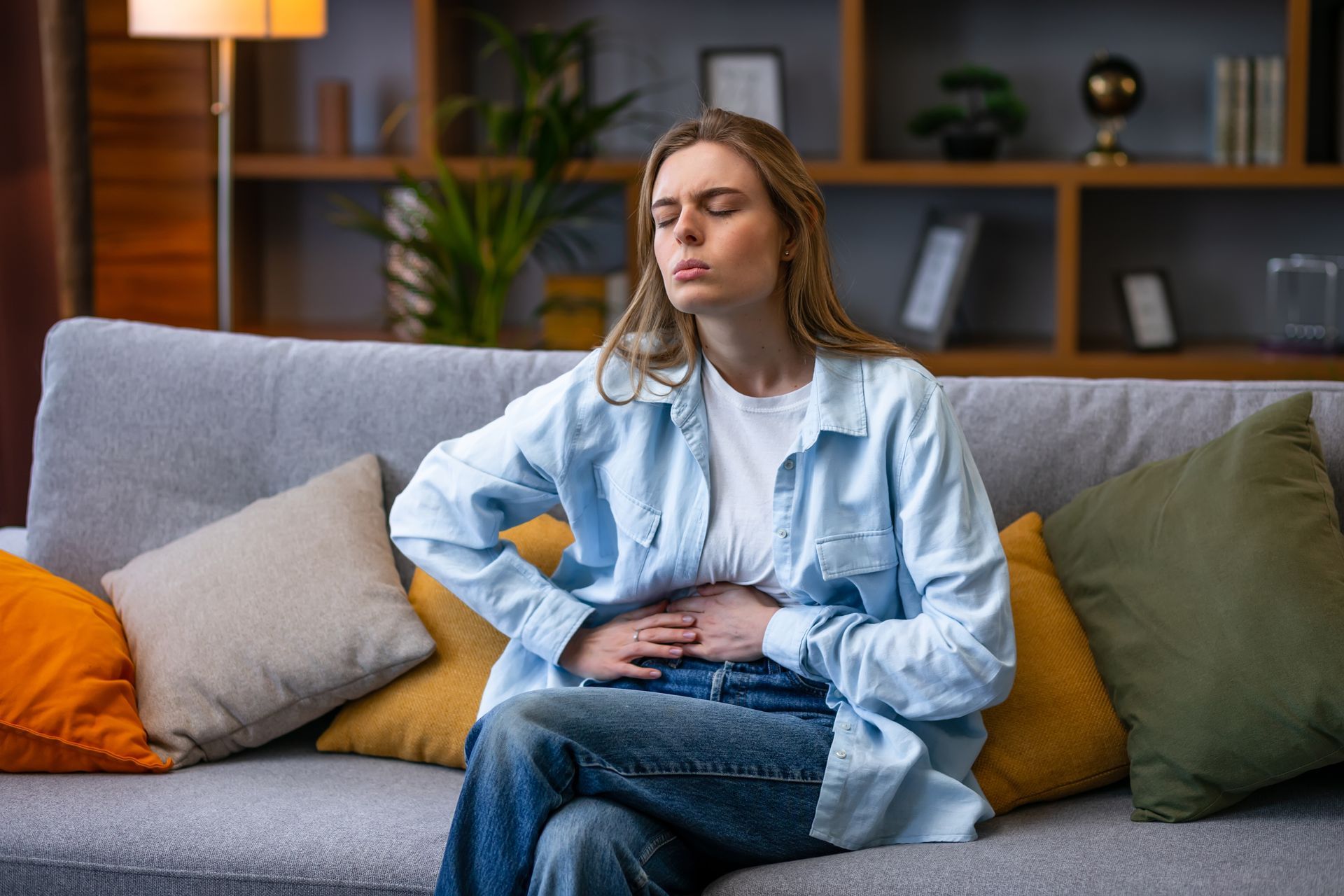 Woman sitting on a couch, clutching her stomach in pain, eyes closed. She's wearing a blue shirt and jeans indoors.
