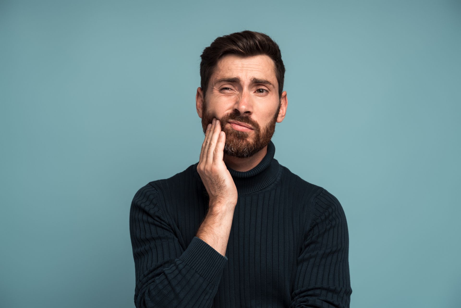 Man with a beard, wearing a dark turtleneck, touching his cheek, showing a pained expression against a blue background.
