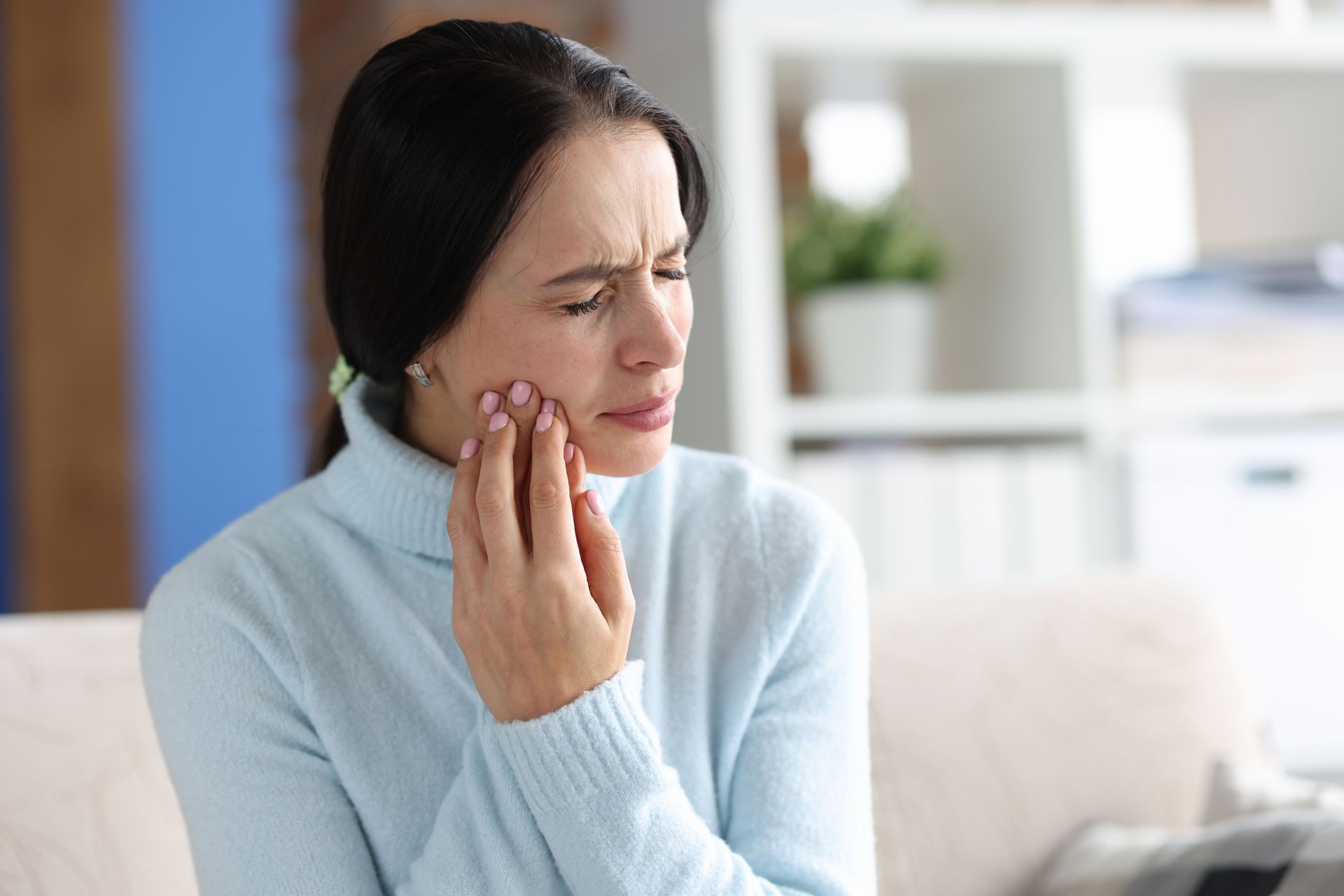 Woman with dark hair in a blue sweater, holding her jaw in pain, indoors.