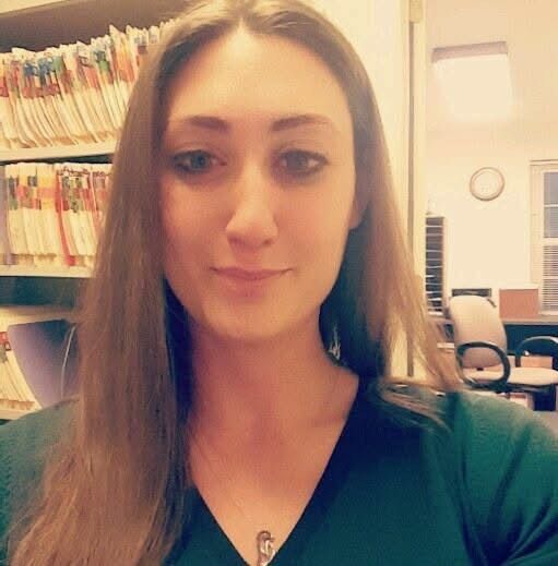 Woman with long brown hair, looking at the camera. She is in an office, with file folders in the background.