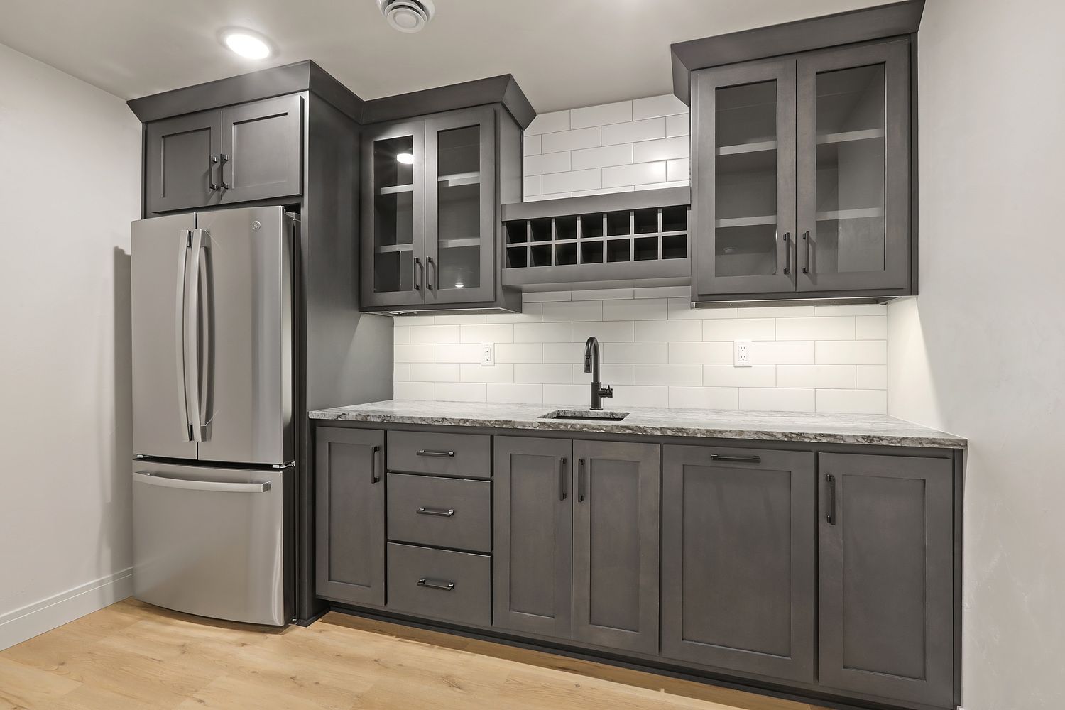 A kitchen with gray cabinets , a stainless steel refrigerator , a sink , and a wine rack.