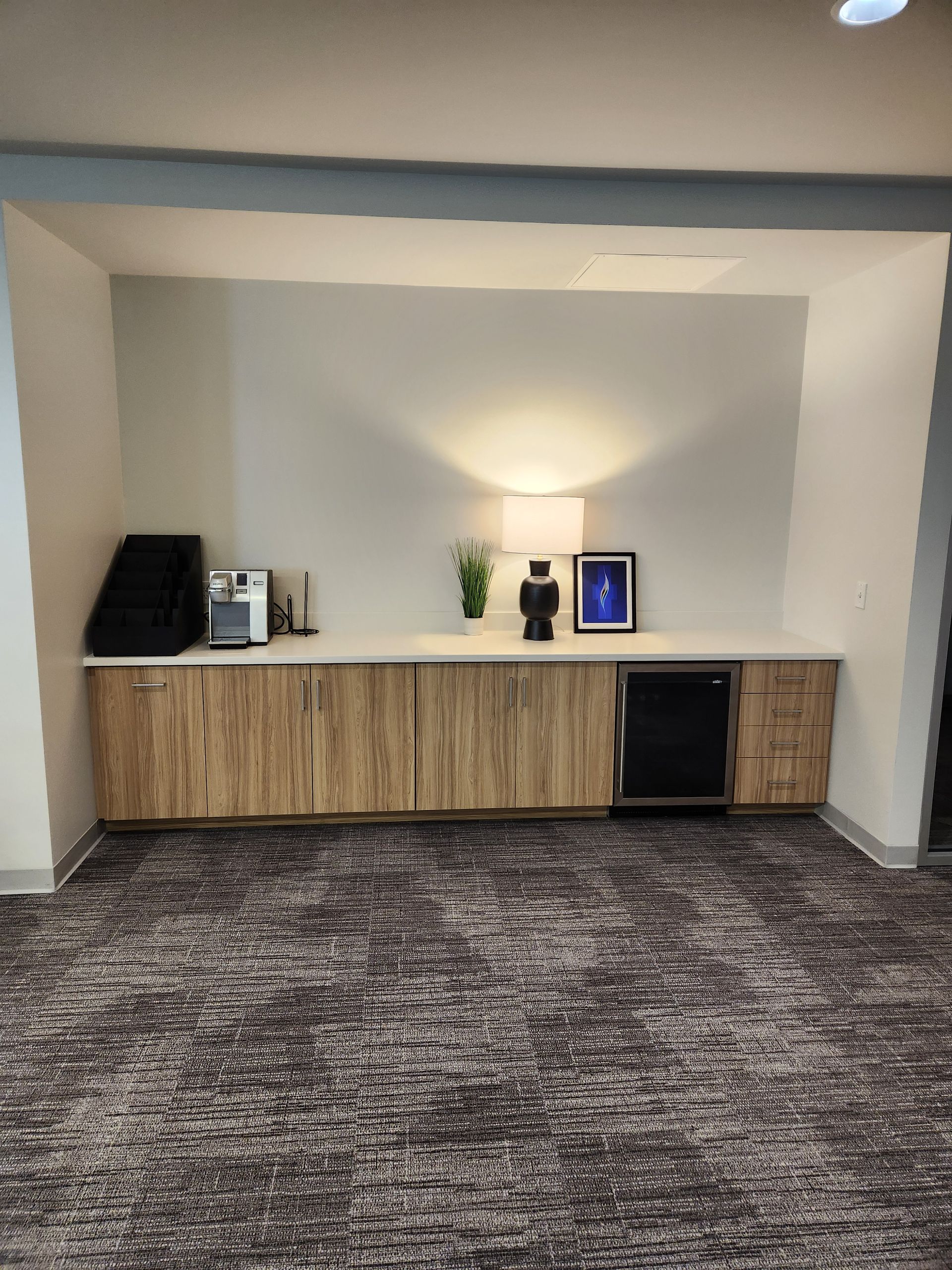 Built-in counter with cabinets and a mini-fridge, with coffee machines, lamp, and decor. Brown carpet in an office setting.