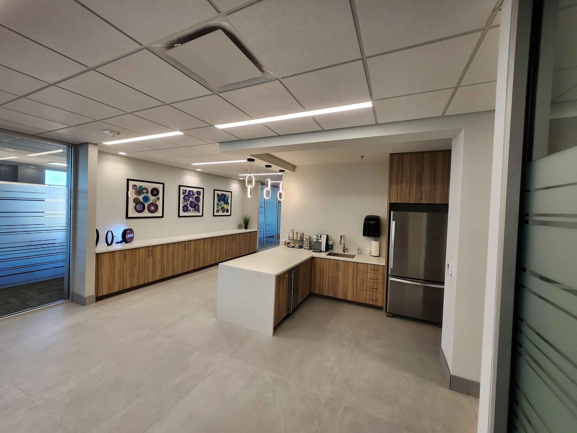 Modern office kitchen with light wood cabinets, stainless steel fridge, and white countertops.