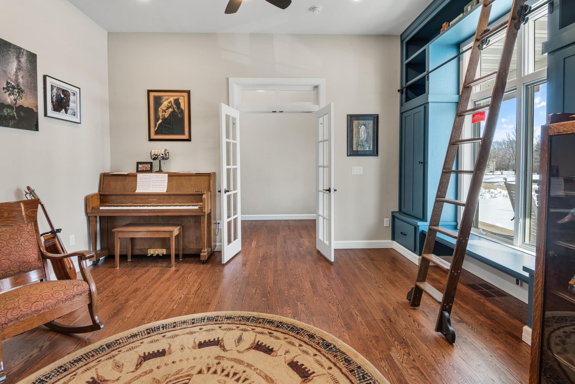 Room with piano, bookcase, and French doors. A wooden ladder leans against a large window.