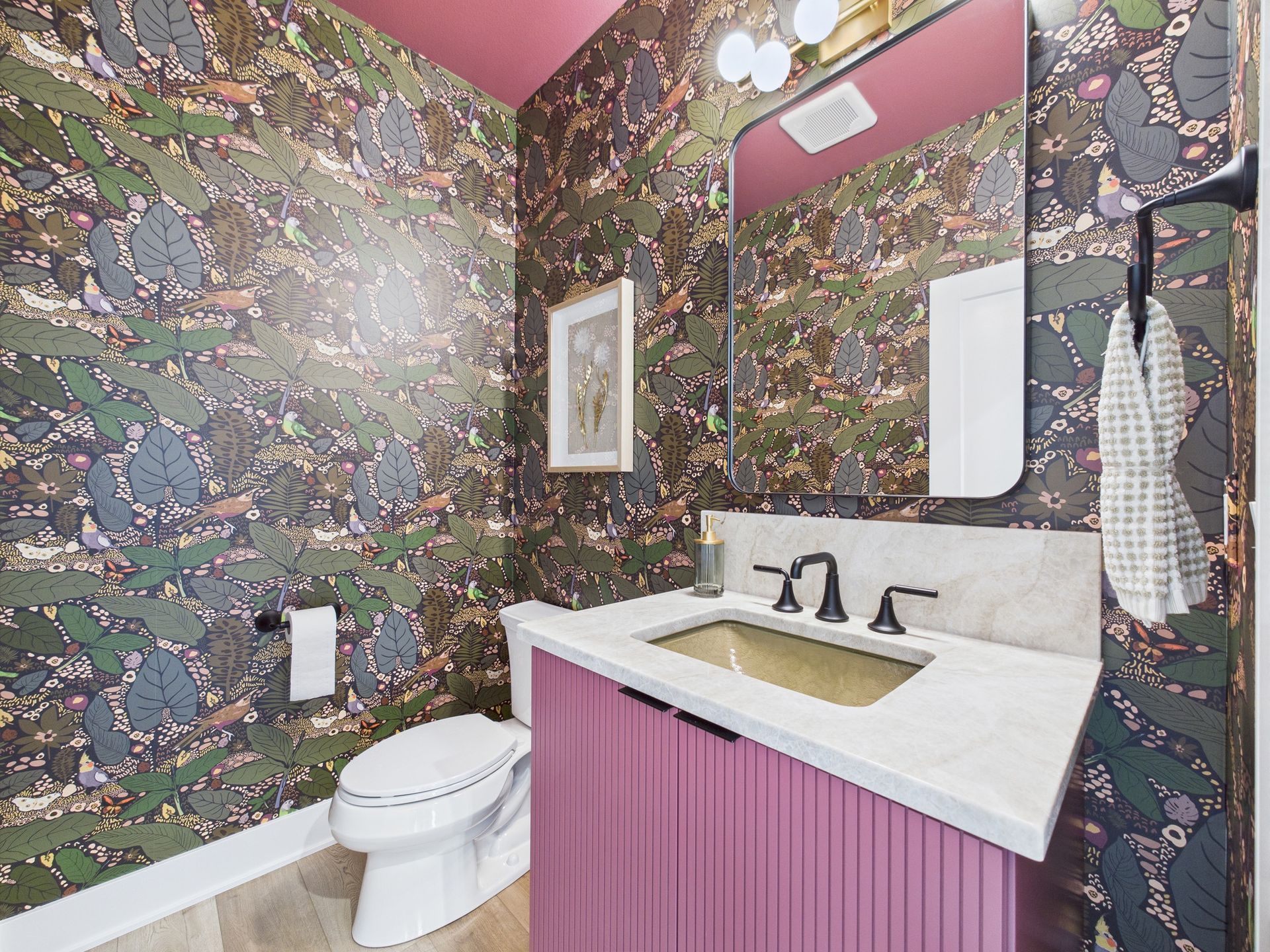 Small bathroom with floral wallpaper, pink vanity, and black fixtures.