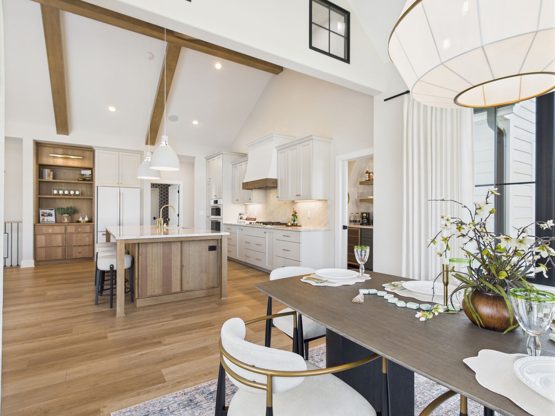 Open-concept kitchen and dining area with wooden beams, island, and table set for a meal. Bright, neutral tones.