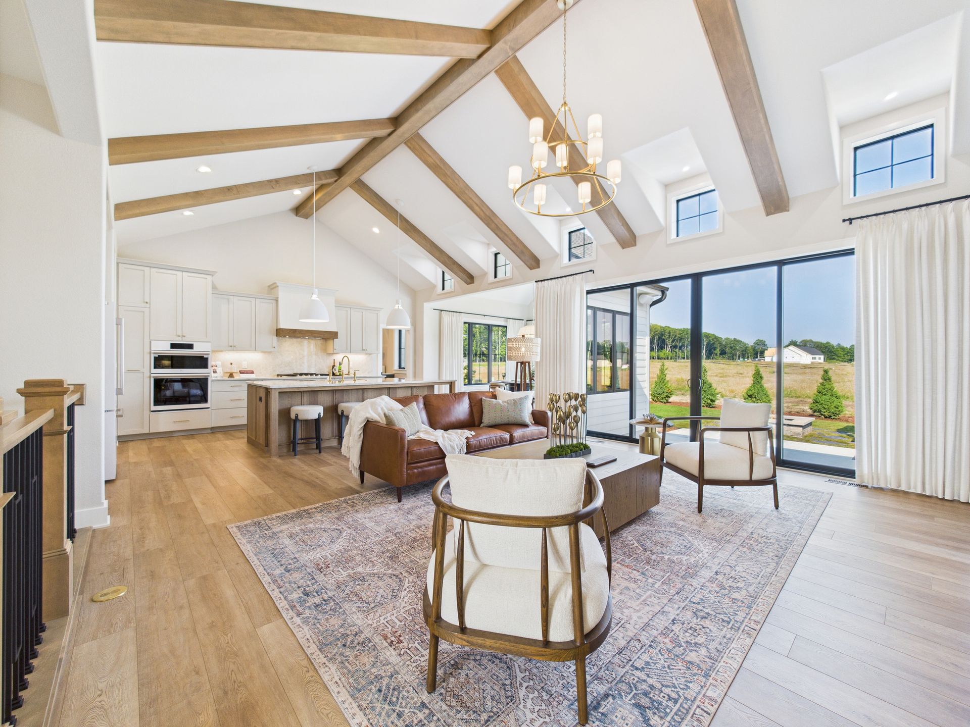 Bright living room with vaulted ceiling, beams, and large windows overlooking a green field.