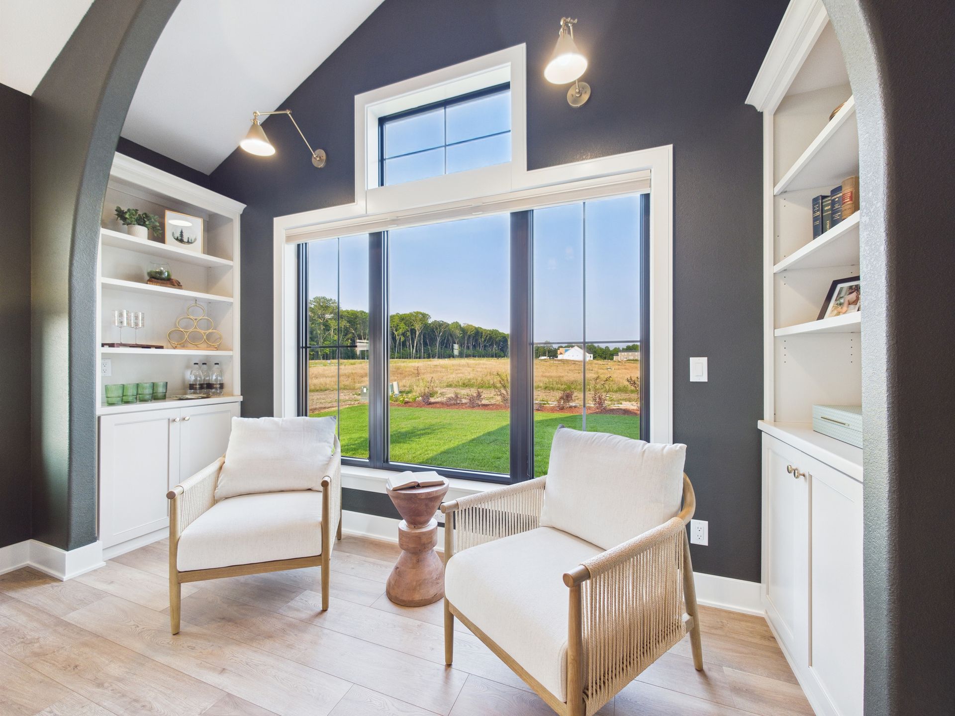 A well-lit reading nook with white armchairs, built-in bookshelves, and a large window overlooking a field.