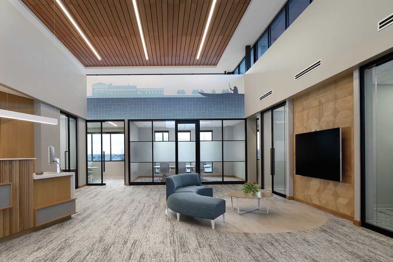 Modern office reception area, blue seating, wood ceiling, glass-walled rooms, neutral tones, large TV screen.