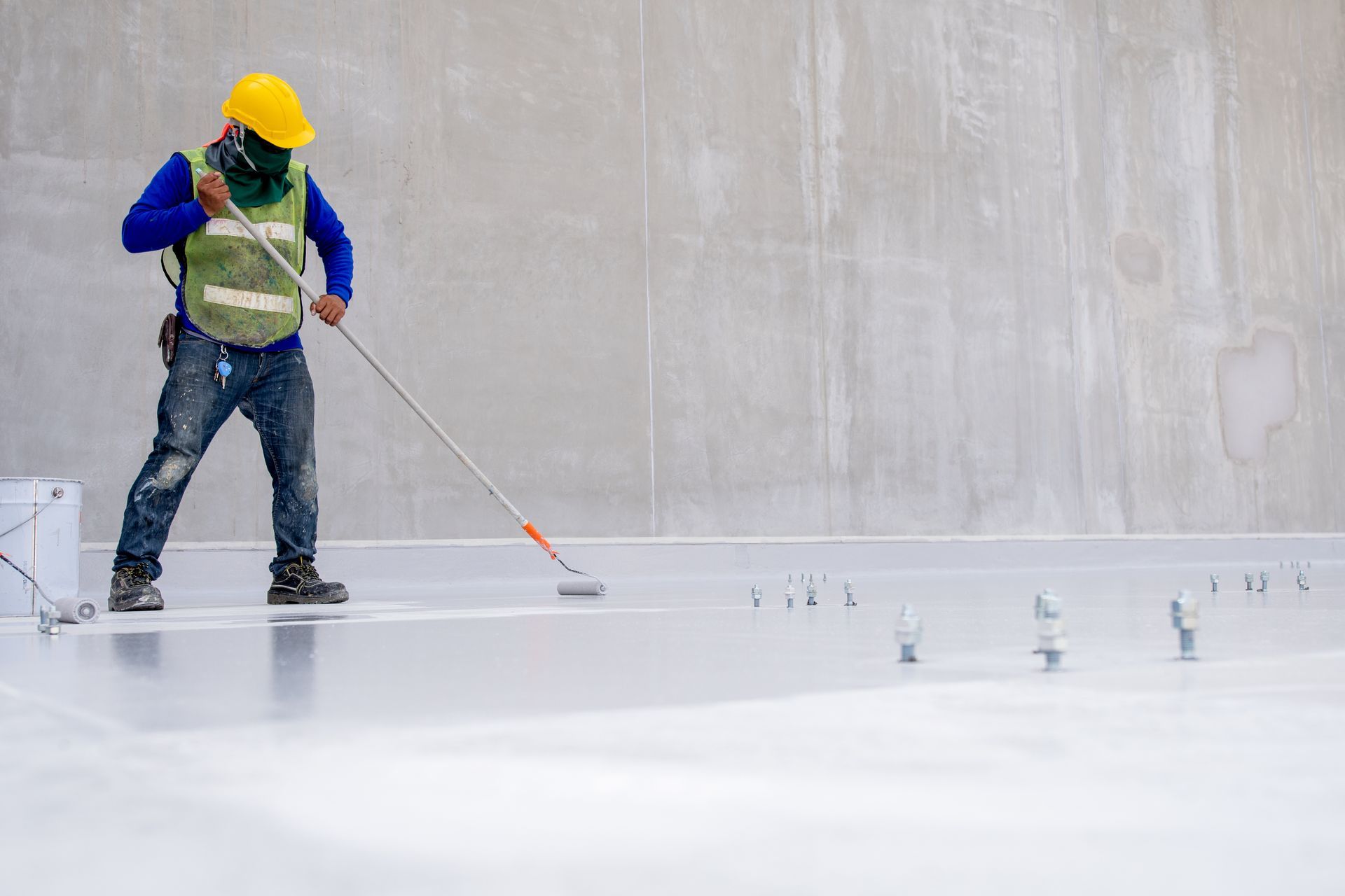 A construction worker is painting a concrete floor with a roller.