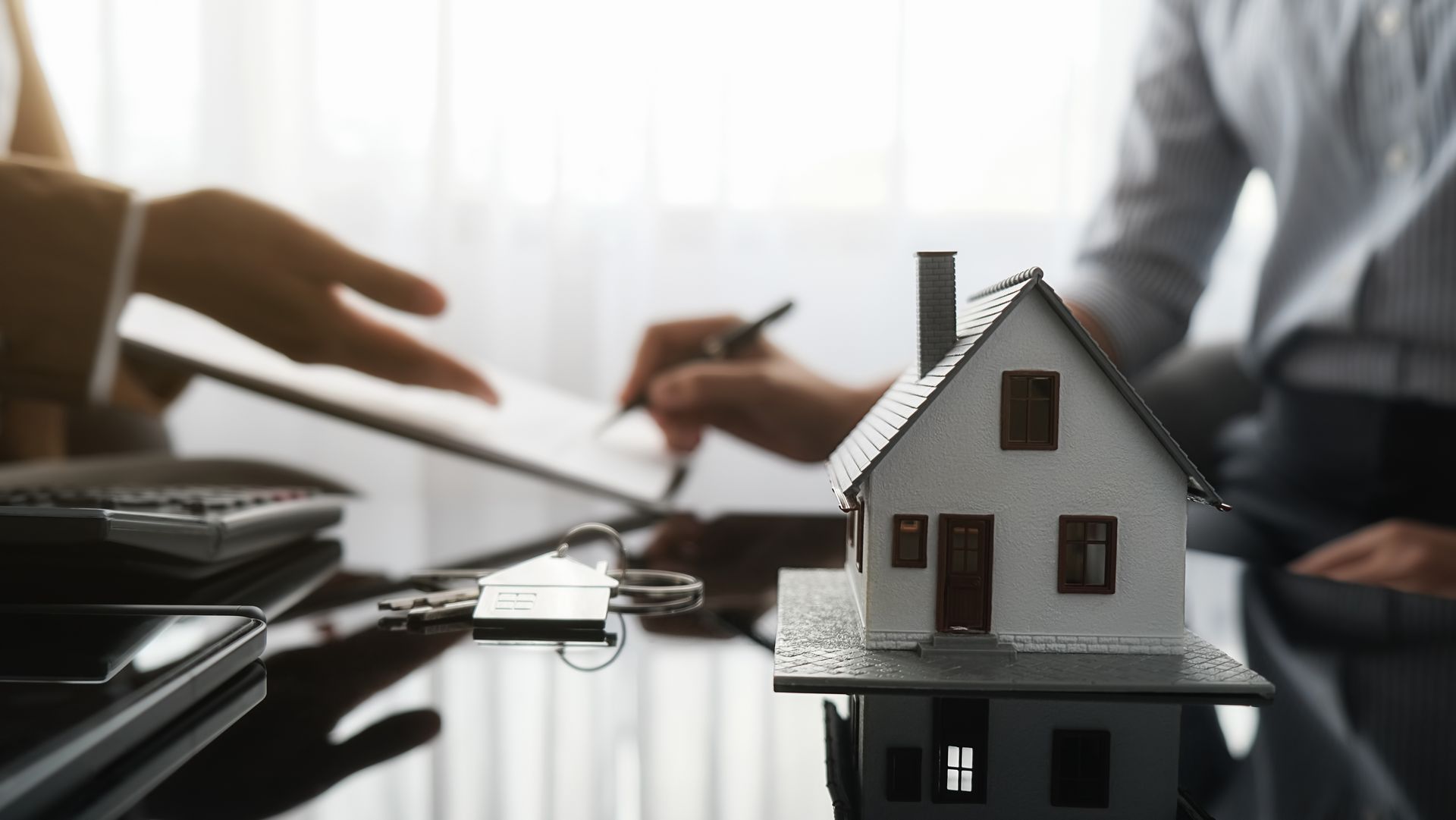Miniature house on a table next to paperwork and keys as someone signs a document.
