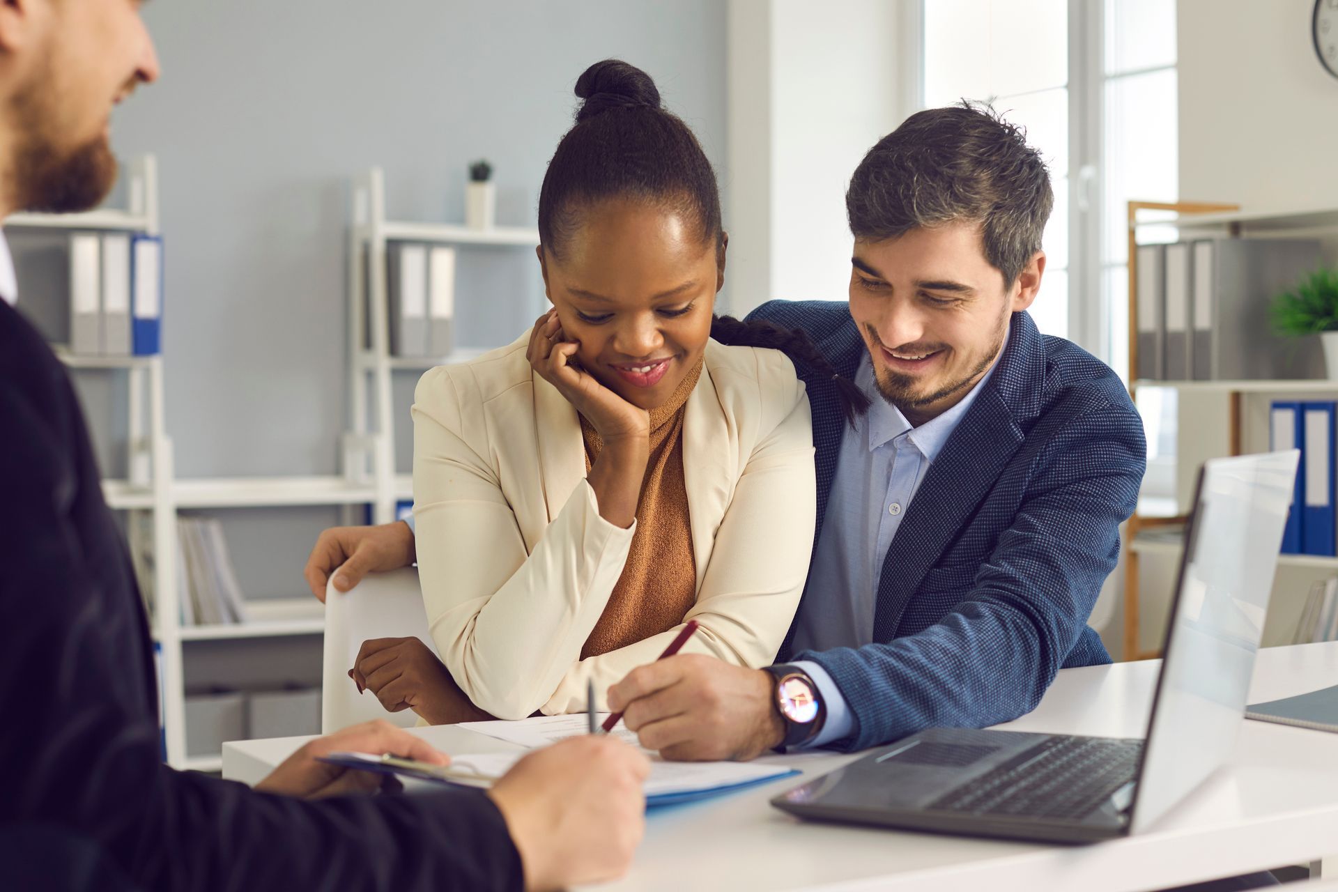 Couple signs paperwork, guided by a person. They appear happy in an office setting.