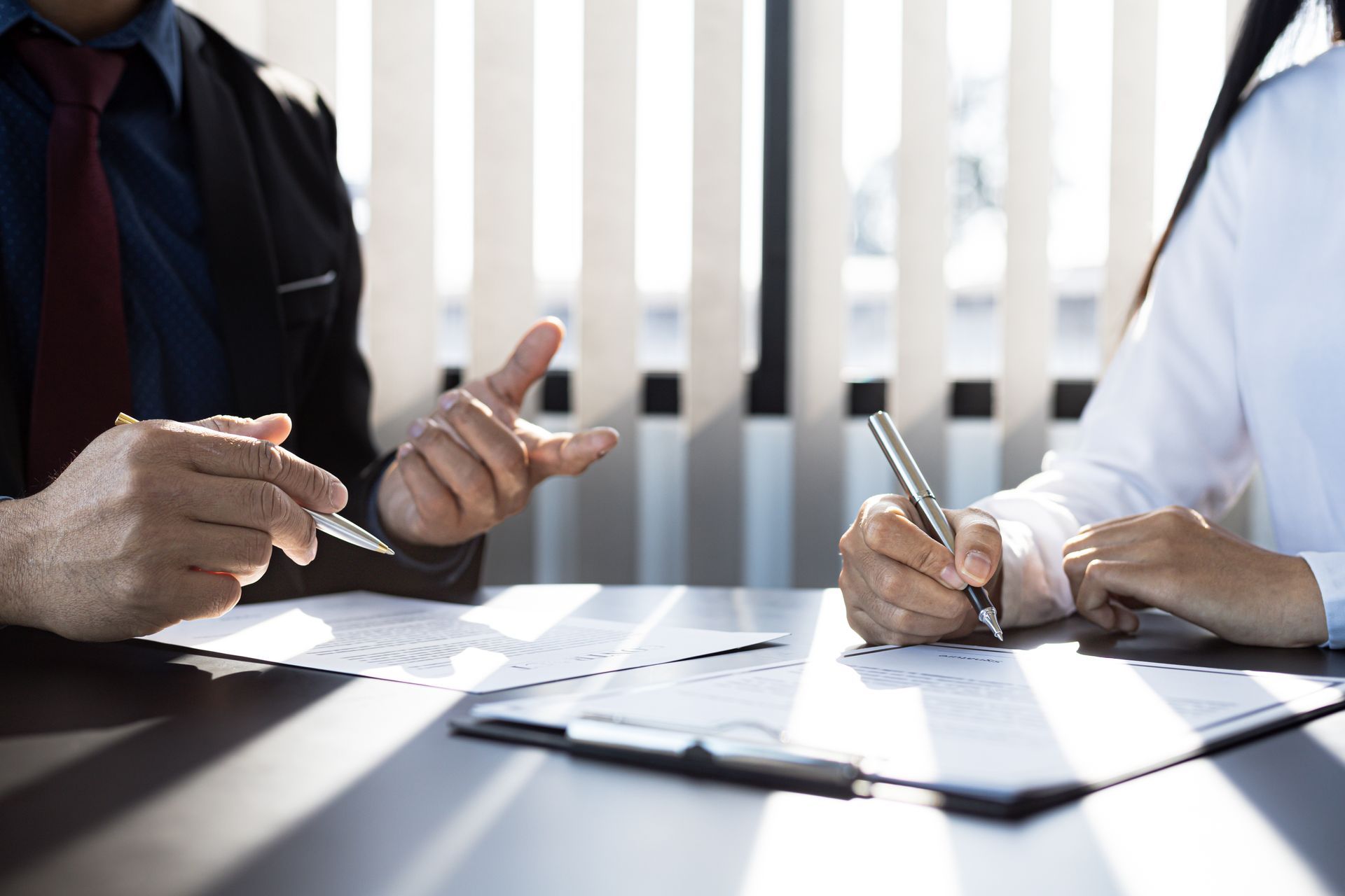 Man gesturing, explaining to person signing document at a table, sunny office setting.