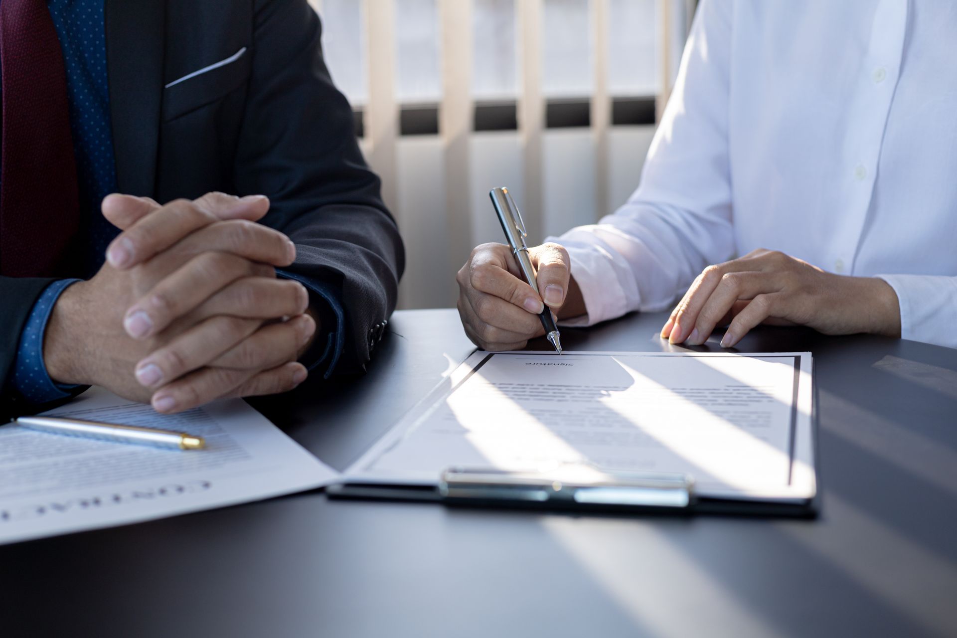 Two people at a desk, one signing a document. A pen rests on the document.