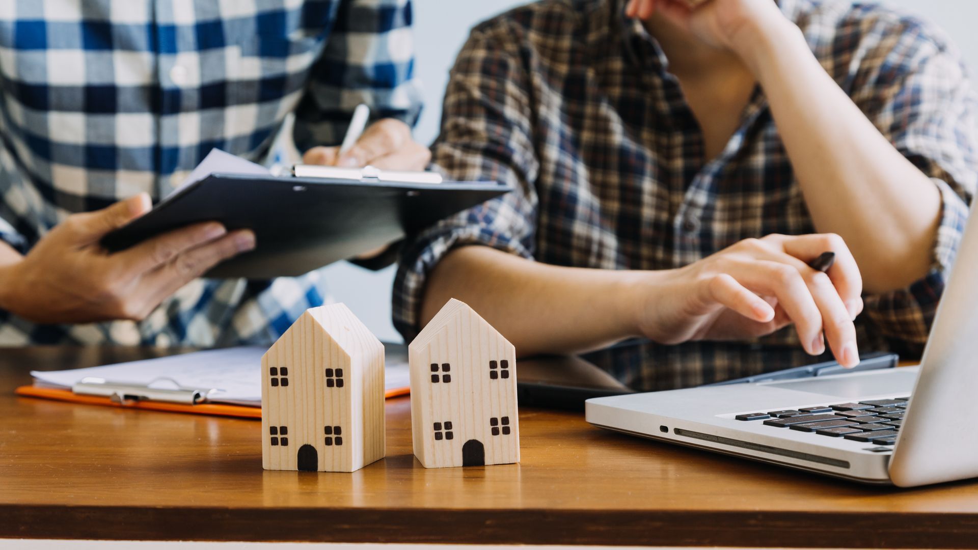 Two people review documents and use a laptop, with wooden house models on the table.
