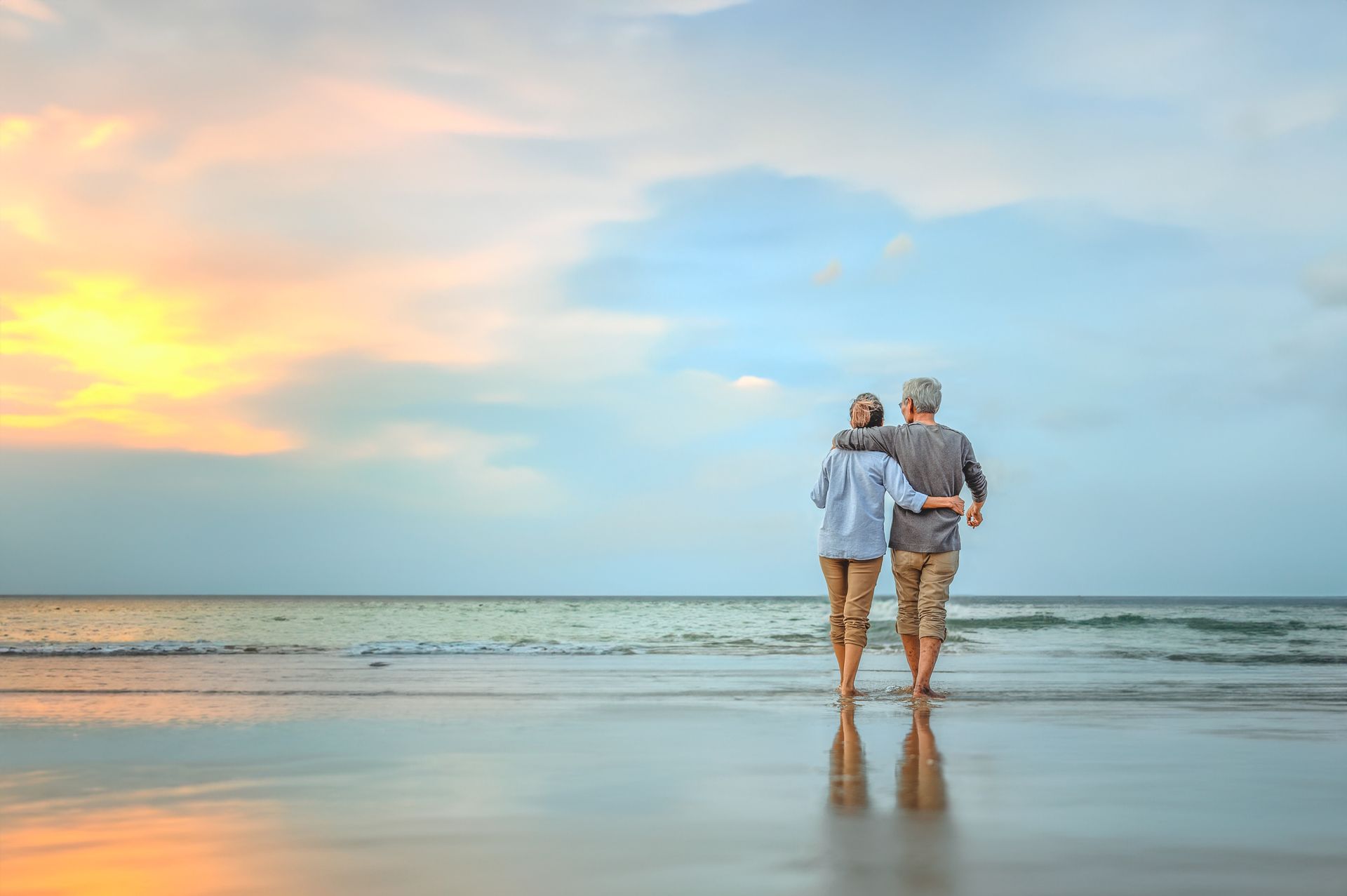 Couple walking on beach at sunset, arms around each other. Reflection in wet sand.