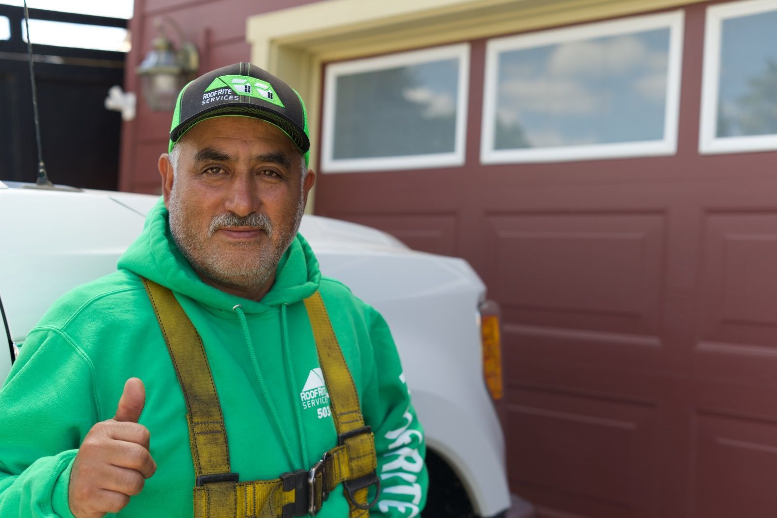 Man in green hoodie and hat gives thumbs up near a truck and garage.