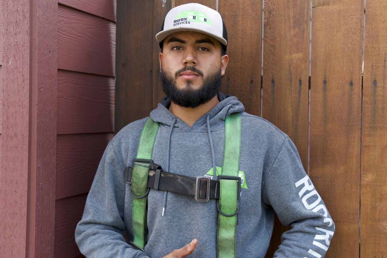 Man in gray hoodie and safety harness, leaning against a wood wall.
